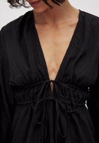 Čierne textúrované šaty s hlbokým výstrihom do V a zhrnutým pásom. Obsahujú dlhé rukávy a zaväzovanie na golieri.