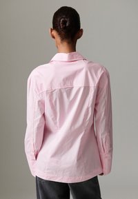 Šviesiai rožinė, ilgomis rankovėmis, su sagomis, marškinėliai su klasikiniais apykakle, tiesia apačia ir nugaros dalies yoke siūlėmis, pagaminti iš lygaus audinio.