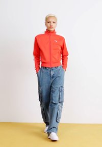 Giacca da track in zip rossa con accenti bianchi e logo, abbinata a pantaloni cargo blu larghi con tasche laterali e sneakers bianche.
