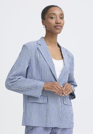 Kobieta ubrana w niebiesko-białą marynarkę w drobną kratkę i pasujące do niej spodnie, z białą bluzką pod spodem, stojąca na jednolitym tle.