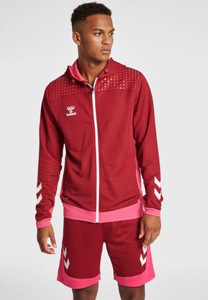 Rode sportjack met ritssluiting, gestippeld patroon, roze accenten en logo, gecombineerd met bijpassende rode shorts met roze bies en chevron-details.