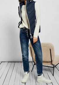 Námořnický modrý péřový vest s bílým zipem a stahovacími šňůrkami, nošený přes smetanový svetr, v kombinaci s modrými džíny a bílými teniskami.