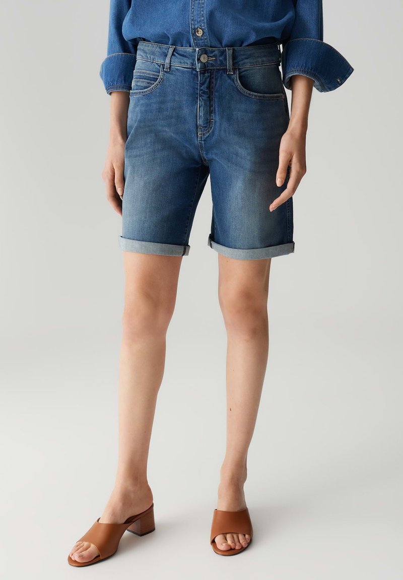 Jeansshorts met een middellange taille, een licht tot donkerblauwe gradiënt, opgerolde zomen en vijf zakken. Gecombineerd met tan instappers met een blokhak.