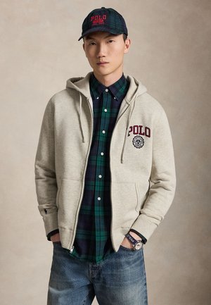 Junger Mann trägt einen grauen Polo-Hoodie, ein grün-navy kariertes Hemd, blaue Jeans, eine Polo-Kappe und eine Armbanduhr mit Polo-Armband.