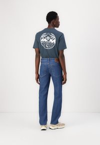 Dark teal T-shirt with a white circular graphic on the back, paired with blue denim jeans and light-colored sneakers. Minimalist design.