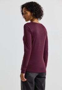 Långärmad burgundy skjorta i ett mjukt, stretchigt tyg med rundad halsringning och figurnära silhuett, parat med mörkgrå byxor.