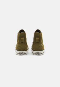 Pár olivově zelených vysokých tenisek Converse All Star zobrazených zezadu na čistém bílém pozadí.