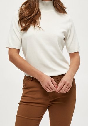 Kobieta w białym golfie z krótkim rękawem i wysokim stanem brązowych spodniach, z rękami złożonymi z przodu.