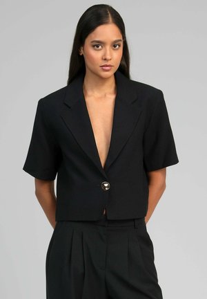 Femme aux longs cheveux foncés porte un blazer noir à manches courtes avec un bouton et un pantalon plissé taille haute assorti sur un fond uni.