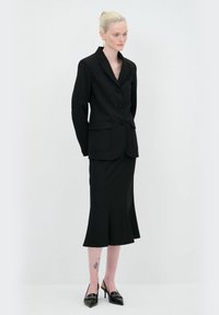 Blazer nero su misura con bottoni, due tasche frontali, abbinato a una gonna aderente fino al ginocchio, tacchi neri a punta affilata e un tatuaggio visibile sulla gamba.