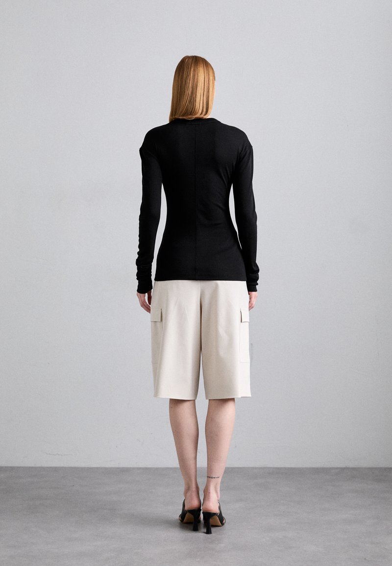 Schwarzes Langarmshirt mit tailliertem Design, kombiniert mit beigen knielangen Shorts, die zwei seitliche Taschen besitzen. Die Schuhe haben einen Blockabsatz.