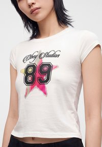 Νεαρή γυναίκα που φοράει λευκό T-shirt με πολύχρωμο αστέρι και μεγάλο γραφικό "89" και μαύρο παντελόνι, σε απλό φόντο.