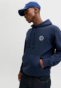 Navy blue hooded sweatshirt with front pocket, featuring a circular logo on the chest. Paired with a matching blue cap. Smooth texture.