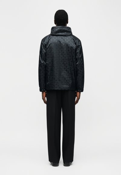 Άτομο που στέκεται με την πλάτη, φορώντας μαύρο μπουφάν με σχέδιο και ψηλό γιακά και φαρδιά μαύρα παντελόνια, μπροστά σε ένα απλό ανοιχτόχρωμο φόντο.