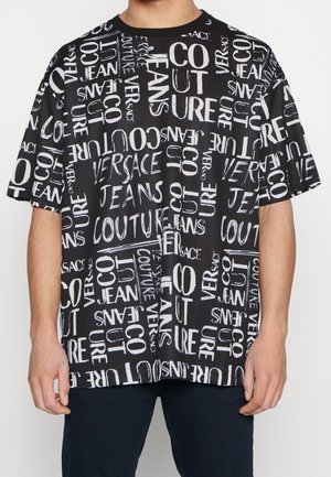Sort T-shirt med korte ærmer med stor hvid tekst, der viser ordene "Versace," "Jeans," "Couture," og "Co" i forskellige skrifttyper og orienteringer.