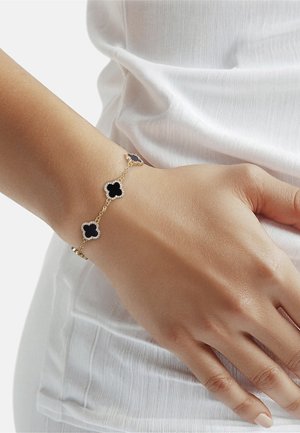 Poignet portant un bracelet en or avec trois breloques en forme de trèfle noir bordées de petites pierres claires, reposant sur un tissu blanc texturé.