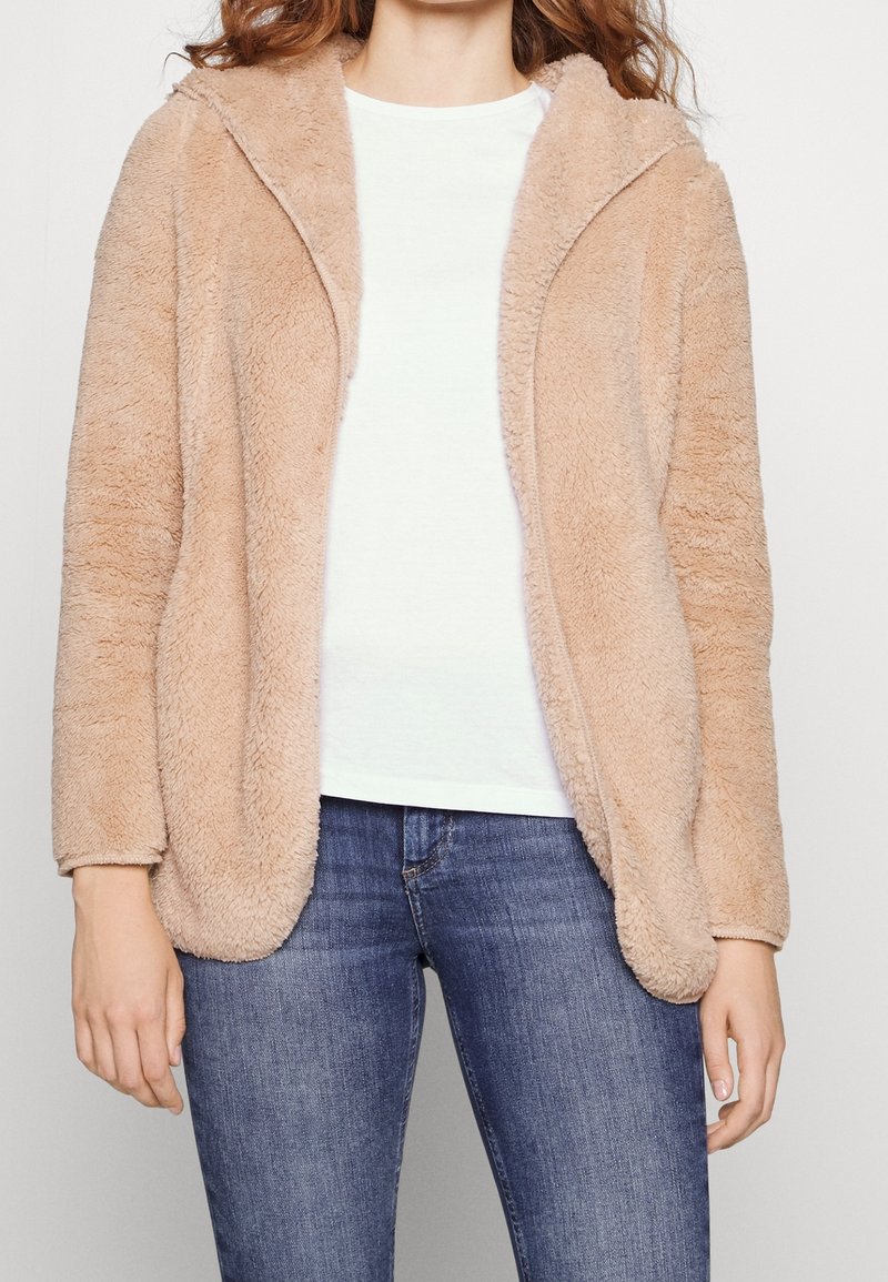 Femme portant une veste à capuche beige en tissu duveteux par-dessus un t-shirt blanc et un jean bleu, debout devant un fond clair uni.