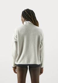 Hellgrauer Fleece-Pullover mit hohem Kragen, sichtbaren Nähten und einem marineblauen Akzent am Saum. Weiche Textur, lockere Passform.