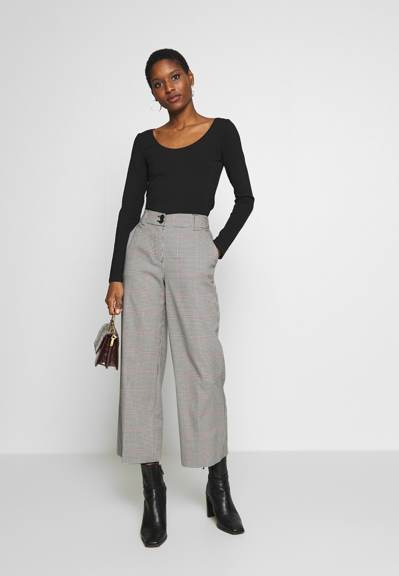Schwarzes Langarmoberteil und hochtaillierte, karierte Hose mit Gürtelakzent. Das Model hält eine kleine Tasche und trägt schwarze Ankle Boots.