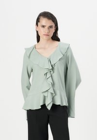Lys grønn bluse med V-hals og store volanger langs fronten og kragen. Lange ermer og en luftig passform.