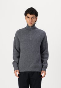 Grauer gerippter Pullover mit hohem Kragen und kurzem Reißverschluss. Strukturiertes Strickmaterial und lange Ärmel. Symmetrisches Design auf der Brust.