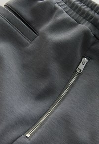 Tissu texturé gris foncé avec une poche zippée argentée et une poche latérale classique sur un pantalon ou un short avec ceinture élastique.