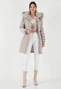 Svijetlosiva puffer jakna s kapuljačom obloženom krznom, uparen s bež argyle puloverom, bijelim hlačama i sjajnim bež čizmama do gležnja.