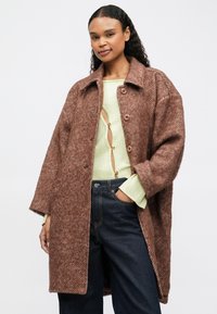 Mulher com cabelo encaracolado, usando um casaco de lã castanho oversized, um suéter canelado verde claro com recortes e jeans azul escuro, posando com a mão no bolso.