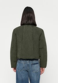 Persoon met schouderlang donker haar, gekleed in een donkergroene gewatteerde jas en blauwe spijkerbroek, met de rug naar een effen witte achtergrond.