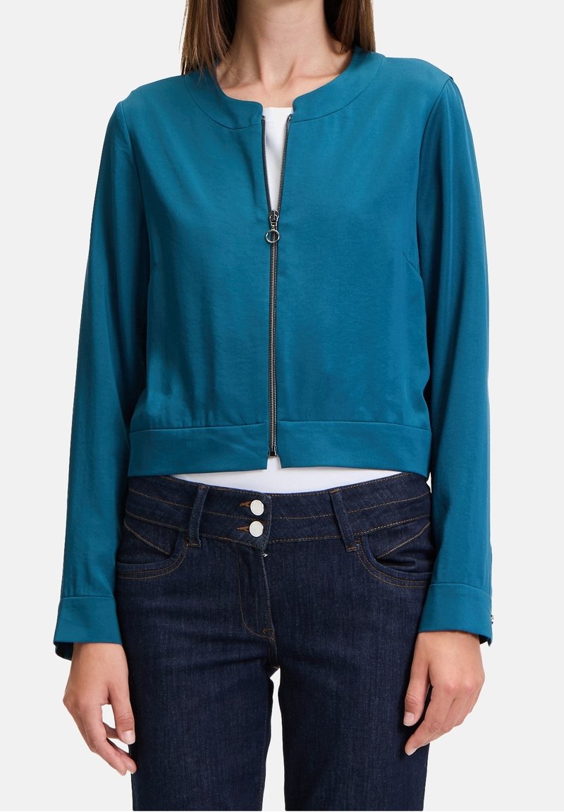 Teal cropped jacket in smooth fabric with a round collar and front zipper. Paired with dark denim jeans featuring double-button closure.