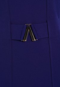 Tissu violet avec une texture lisse, comportant un accent en métal noir et or en forme de "A" à la taille. Détail de ceinture ajustable.