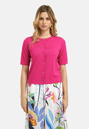 Woman wearing a bright pink short-sleeve buttoned cardigan paired with a white skirt featuring colorful floral and leaf patterns.