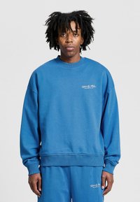 Niebieska bluza z okrągłym dekoltem wykonana z miękkiej mieszanki bawełny, z ribbowanymi mankietami i dołem, z małym białym logo na piersi oraz luźnym krojem.