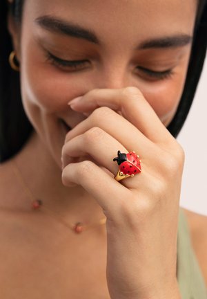 Mujer sonriente con los ojos cerrados, con un anillo dorado en forma de mariquita roja y negra en el dedo índice.