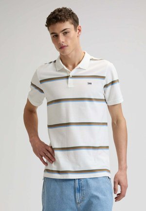 Junger Mann, der ein weißes Poloshirt mit horizontalen blauen und braunen Streifen sowie hellblaue Jeans trägt, vor schlichtem Hintergrund stehend.