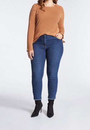 Femme portant un haut marron à manches longues, un jean skinny taille haute bleu avec des ourlets effilochés, et des bottines noires à talons, debout avec une main dans la poche.