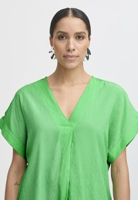 Blusa verde a maniche corte con scollo a V, realizzata in un tessuto leggero e testurizzato. Caratterizzata da una drappeggiatura ariosa e un colore vivace.