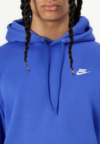 Sweatshirt com capuz azul da Nike, com cordão no capuz e logo branco da Nike no peito. O tecido parece macio e texturizado.