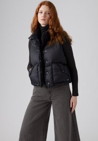 Zwarte puffer vest met een hoge kraag, knopen aan de voorkant en twee grote zakken. Draag het over een zwarte turtleneck, gecombineerd met brede grijze pantalon.