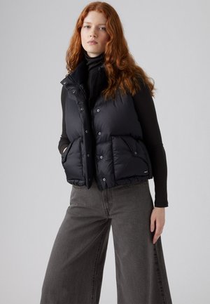 Zwarte puffer vest met een hoge kraag, knopen aan de voorkant en twee grote zakken. Draag het over een zwarte turtleneck, gecombineerd met brede grijze pantalon.