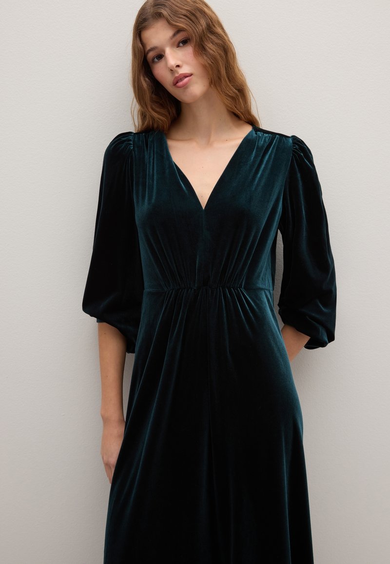 Robe en velours à manches longues de couleur vert profond, avec un col en V et une taille froncée, présentant une texture douce et un drapé fluide.