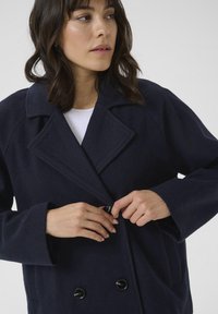 Tamsiai mėlynas dvigubas vilnos mišinys paltas su iškiliais atlapais, dviem priekinėm kišenėm ir juodais mygtukais. Minkšta tekstūra, laisvos formos dizainas.