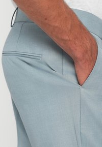 Pantalons ajustés bleu clair avec une texture lisse, présentant une poche latérale et une poche arrière avec un léger détail de rabat.