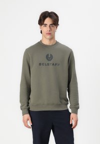 Maslinasto zelena dukserica s okruglim izrezom i rebrastim manžetama. Ispisani "BELSTAFF" logo u tamno sivoj boji preko prsnog dijela. Glatka tekstura.