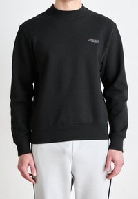 Černá mikina s kulatým výstřihem a malým logem "BOSS" na hrudi, oblečená s světle šedými kalhotami s černými bočními pruhy.