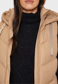 Beige Puffer-Weste mit Kapuze, ausgestattet mit einem Reißverschluss vorne, Seitentaschen und einem strukturierten schwarzen Pullover darunter. Glattes Nylonmaterial.