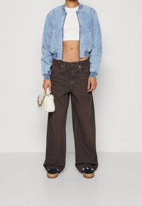 Džínová bomber bunda světle modré barvy, bílý crop top, hnědé kalhoty s širokými nohavicemi a viditelným šitím, a malá bílá texturovaná kabelka.