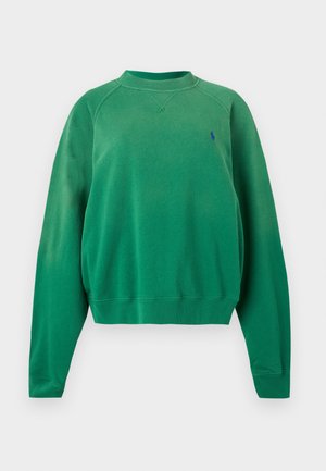 Γυναίκα με πράσινο φούτερ, μπλε τζιν, λευκά αθλητικά παπούτσια, κρατώντας μικρή καφέ τσάντα, στέκεται μπροστά σε απλό ανοιχτό φόντο.