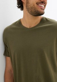 Uomo con capelli corti e ricci e barba che sorride, indossando una maglietta a V verde oliva su uno sfondo bianco.