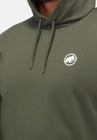 Uomo che indossa un felpa verde oliva con cappuccio e cordini, caratterizzata da un piccolo logo di mammuth bianco sul petto sinistro.